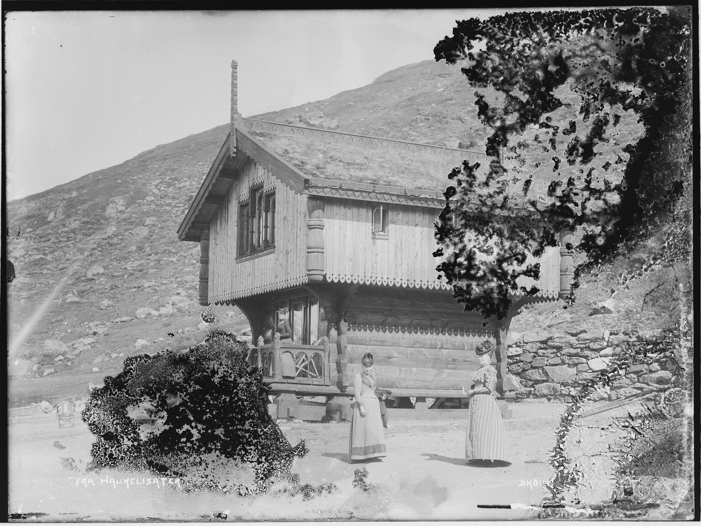A black-and-white photograph of a timber storehouse and two women, with black branching mold across the roof and the foreground
