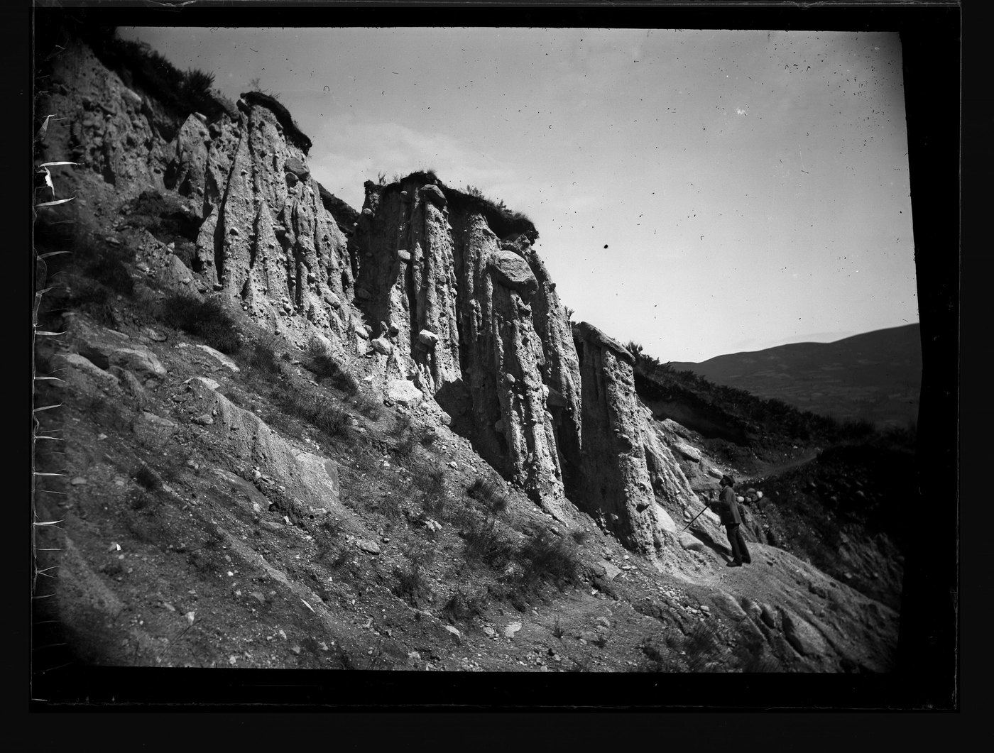 A glass plate photograph of an eroded hillside with a standing figure, its corners darkened and a black wedge at the right edge