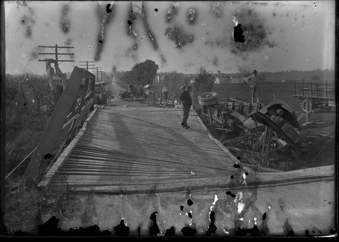 A black-and-white photograph of men and machinery at a timber bridge, the emulsion blistered along the top and bottom edges
